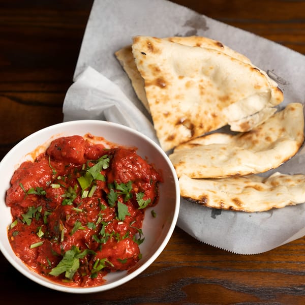 A white bowl of red Indian curry with cilantro, next to a basket of naan bread.