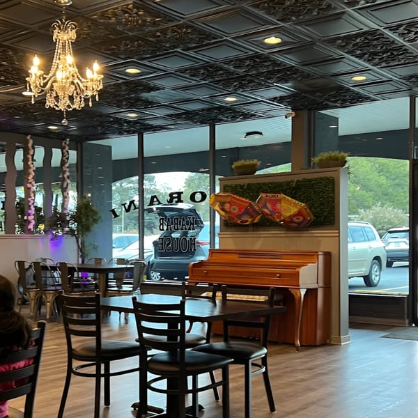 Interior of Noorani Kabab House with tables, chairs, a chandelier, and a piano near the window.