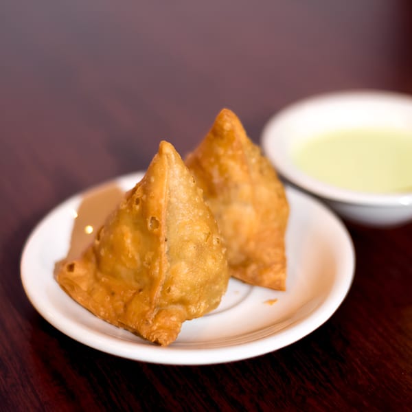 Two golden-brown samosas on a white plate with a small bowl of light green sauce.
